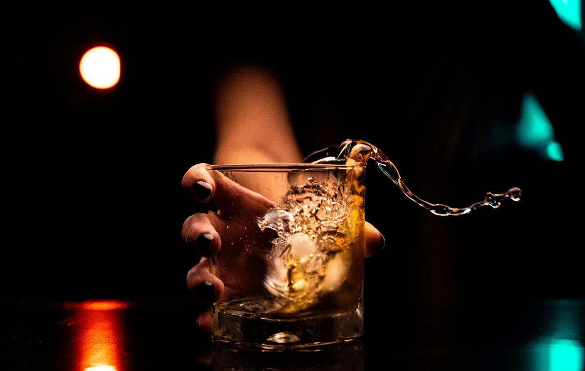 Person holding a glass of whiskey with liquid splashing over ice, symbolizing the struggle to control drinking—representing online alcohol rehab support at Freeman Recovery Online.