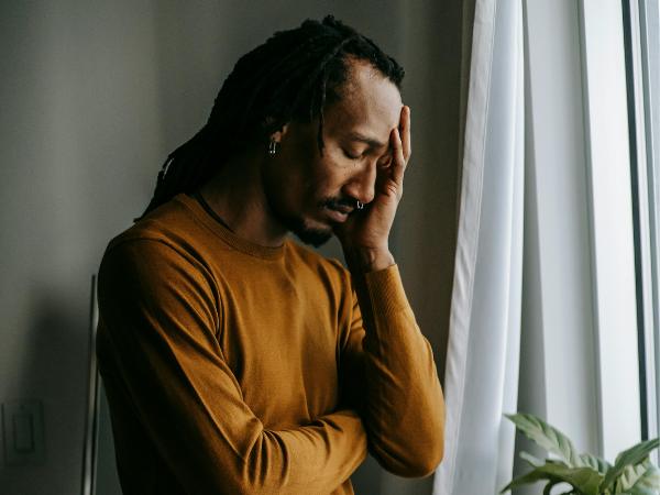 An anxious man standing next to a window with his head down and left hand on his head