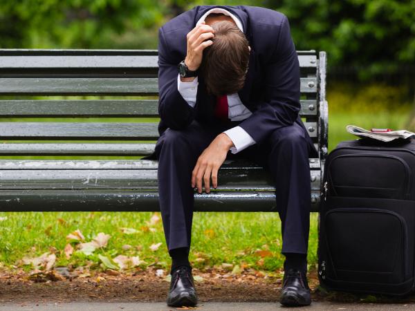 A depressed businessman sits on a park bench with his head down and a black luggage bad next to him