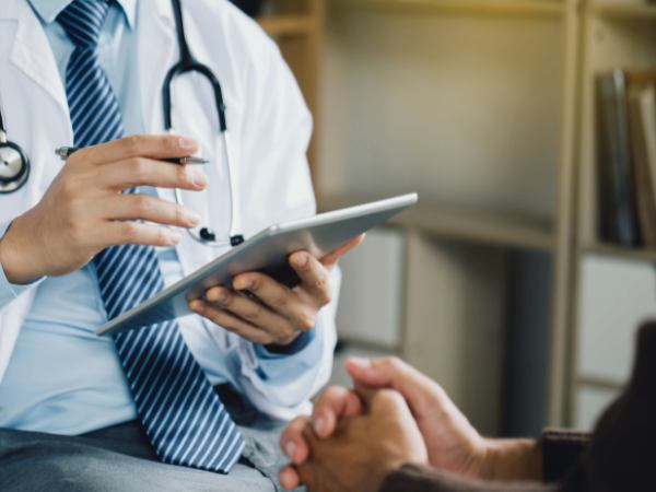 A below-the-shoulder view of a medical professional with a tablet having a mental health consultation with a patient whose hands-only are in the picture