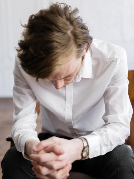 A young man wearing a business shirt, experiencing addiction fatigue and sits with his hands folded and his head down