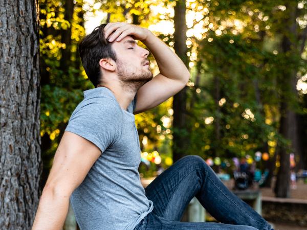 Man outdoors sitting against a tree and holding his head to to substance addiction
