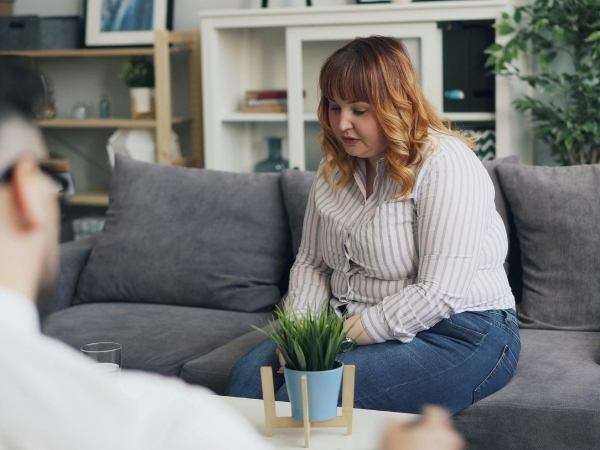 A woman sitting on a couch and talking with her head down as a mental health therapist takes notes