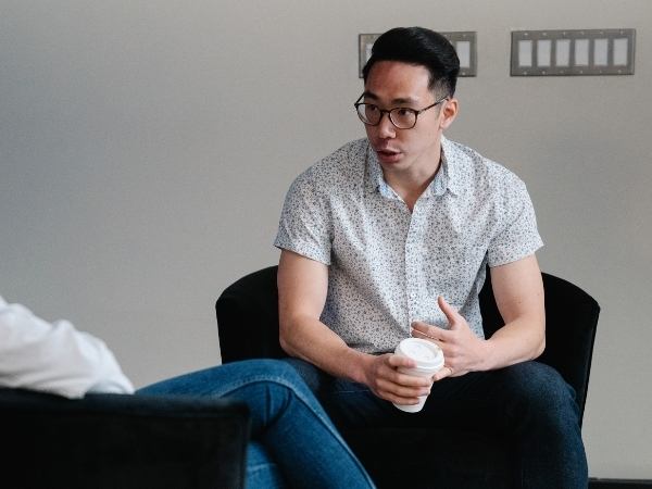 A young man holding a cup of coffee while talking to a mental health therapist who is only visible from a shoulder-to-calf side view