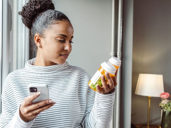 A young woman on a medication management video call, holding her phone in one hand and two prescription medication bottles in the other hand