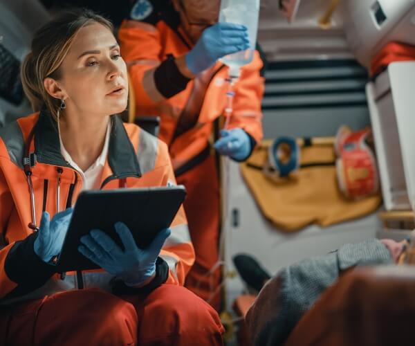 A first responder takes notes on a tablet as she speaks with a patient while her coworker prepares an IV bag